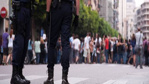 Two police officers stand guard facing a crowd of people on a city street. The officers are wearing dark uniforms and boots, with their backs to the camera.